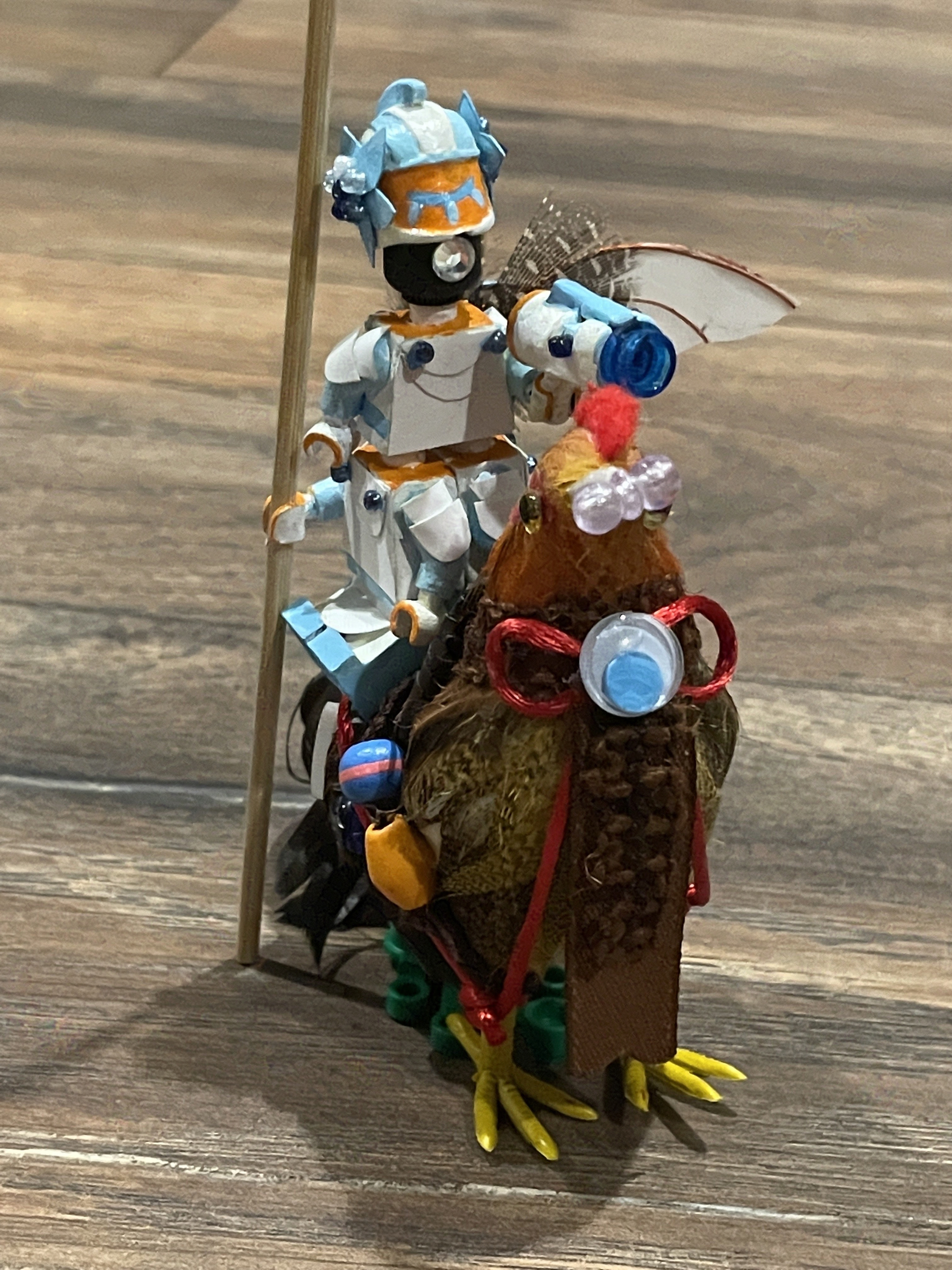 Three lego figures stacked to form a single man, riding a chicken. The man's head is pitch black with a single white rhinestone eye. He is wearing white and blue paper armor. The chicken has a bridle and a googly-eye bowtie.