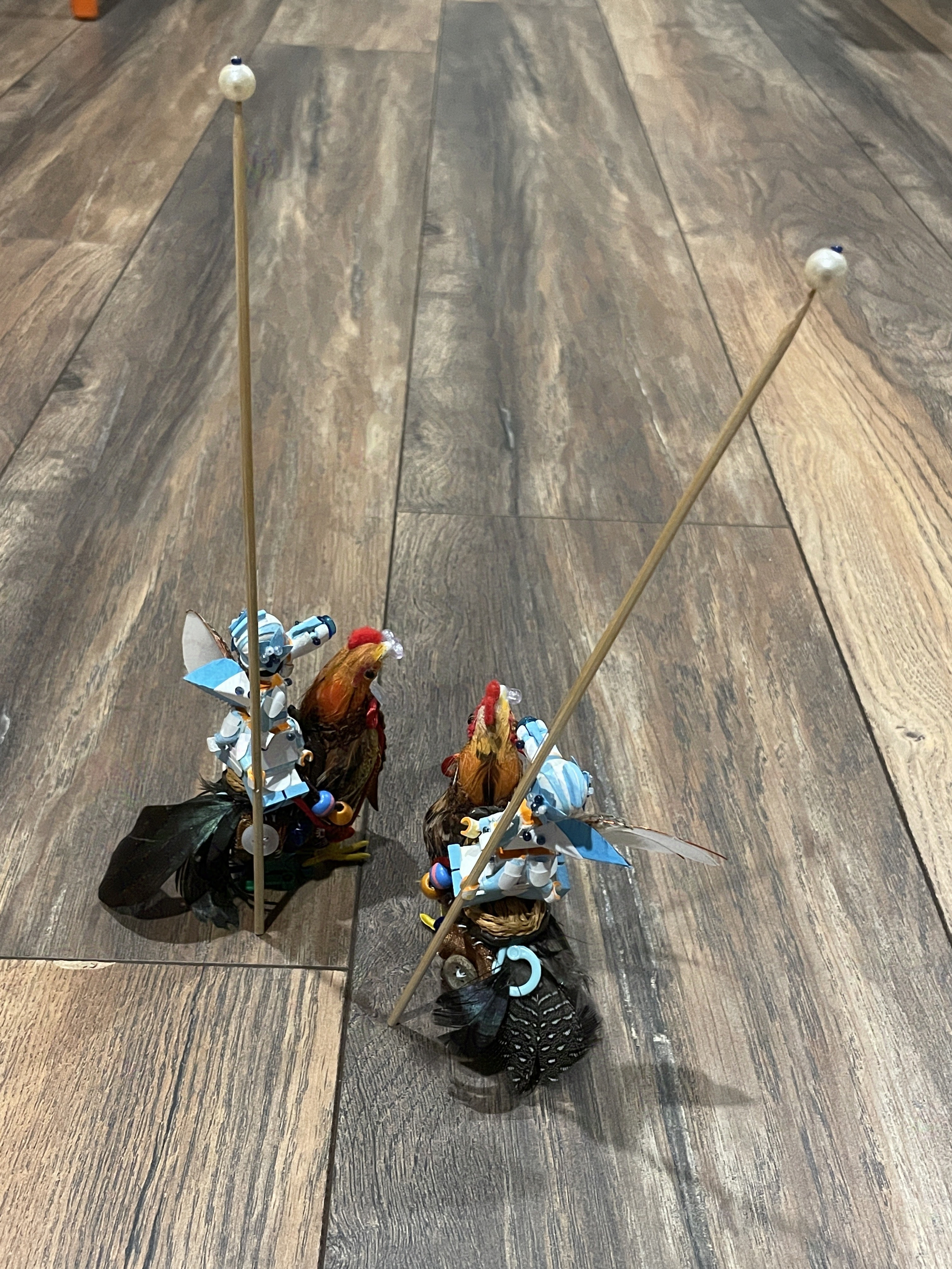 Two identical lego men riding separate chickens on a wood floor. They have pearl-tipped wooden staves that are over double their height.