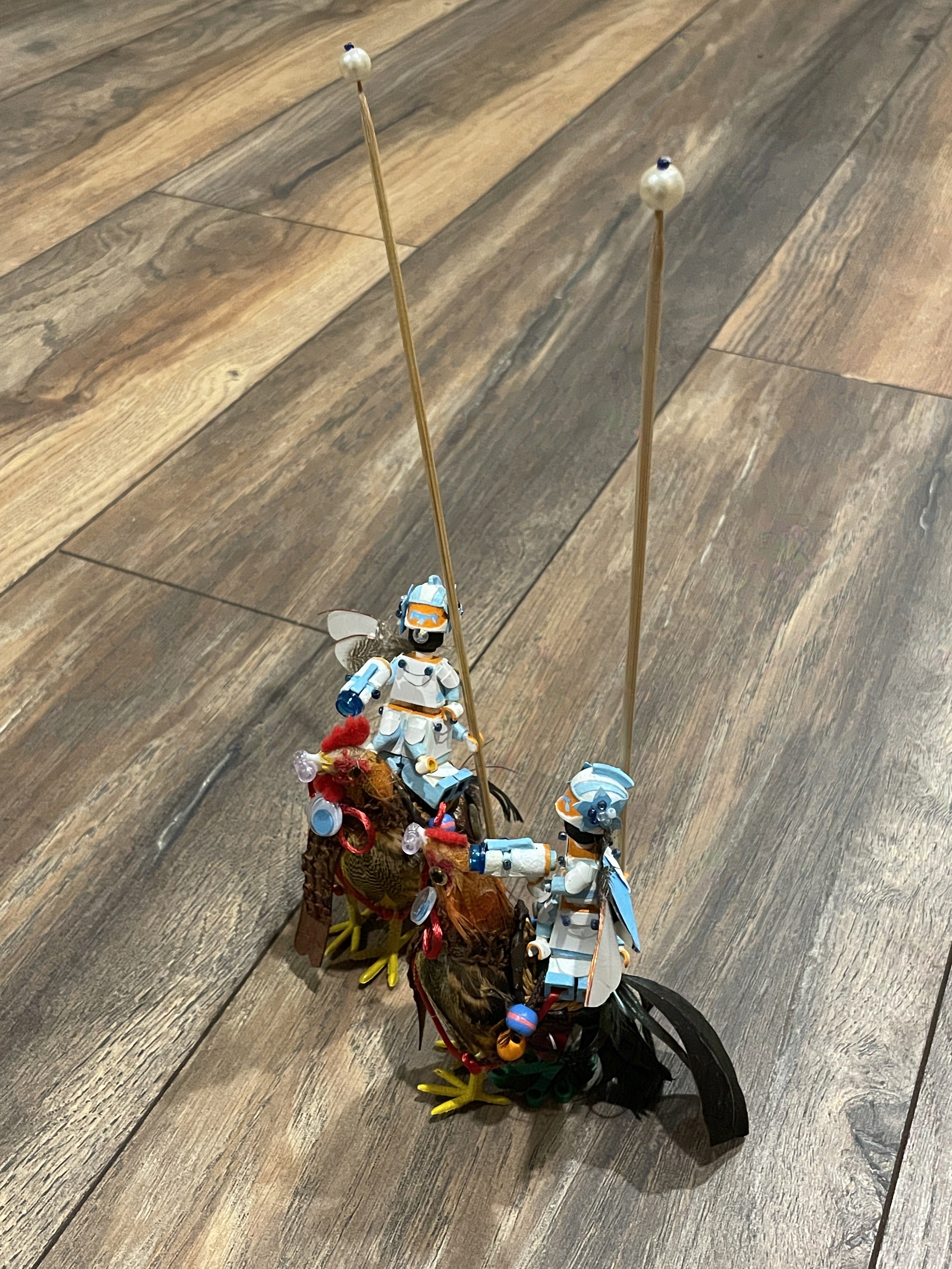 Two identical lego men riding separate chickens on a wood floor. They have pearl-tipped wooden staves that are over double their height.